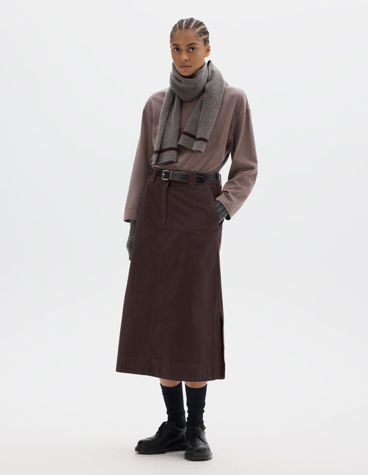 Full body image, front view of model wearing dark brown A line skirt with side splits, double hook and bar fastening waistband, belt loops and slant pockets. Styled with lilac long sleeve top tucked in, grey knitted scarf, black leather belt, black socks and black leather shoes.