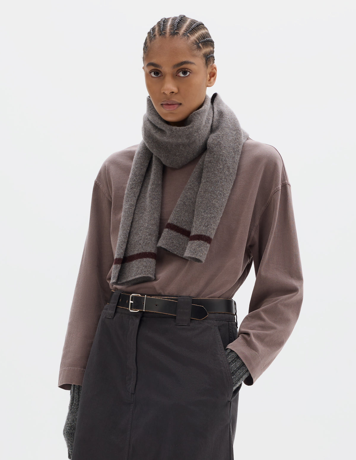Cropped body image, front view of model wearing a brownish grey lambswool scarf with a dark brown stripe, styled with a lilac long sleeve top tucked into a dark brown skirt, black leather belt, and grey wool gloves.