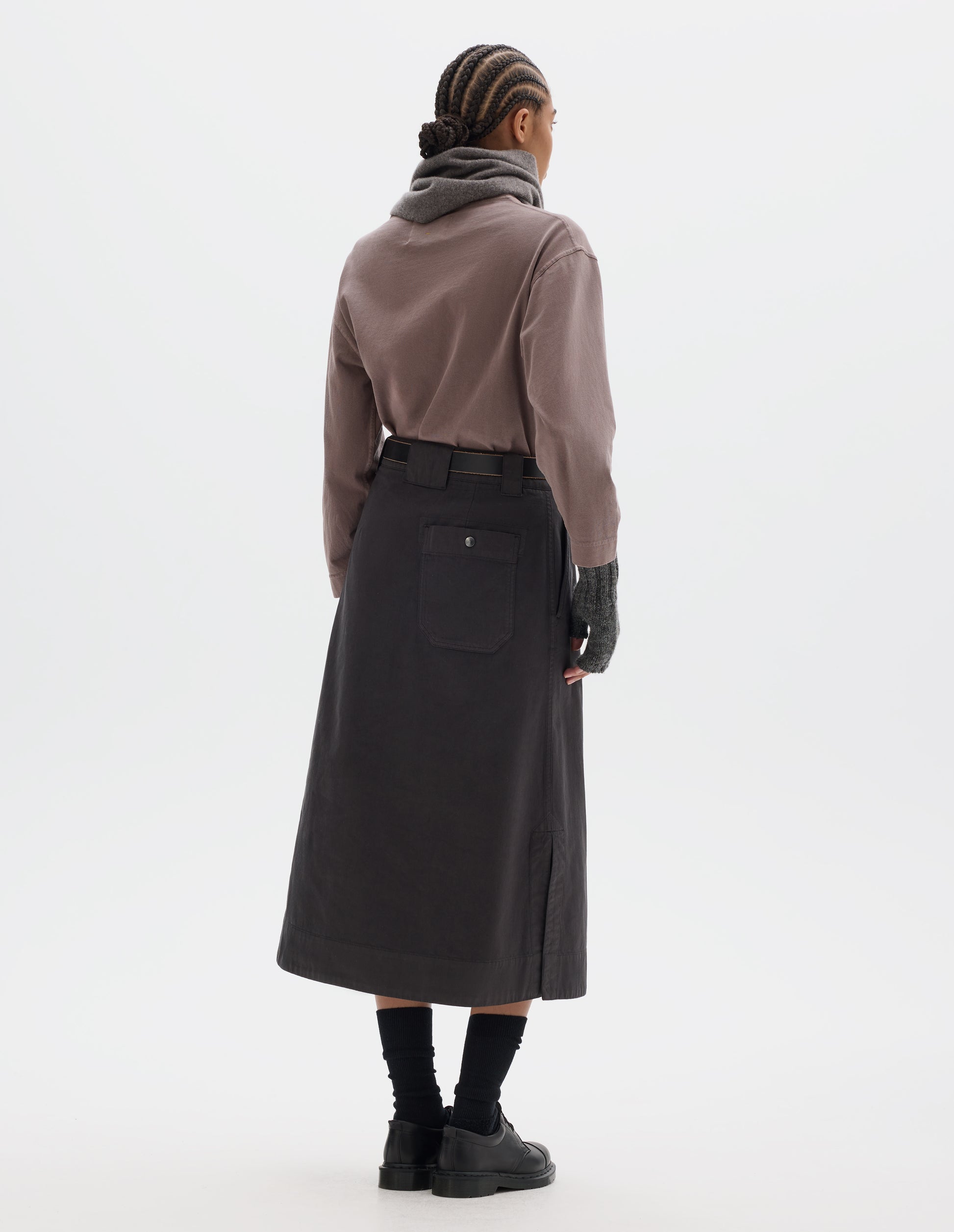 Full body image, back view of model wearing dark brown A line skirt with side splits, belt loop detail on back and back patch pocket with reinforced stitch detailing. Styled with lilac long sleeve top tucked in, black leather belt, grey scarf and matching fingerless gloves. With black socks and black leather shoes.
