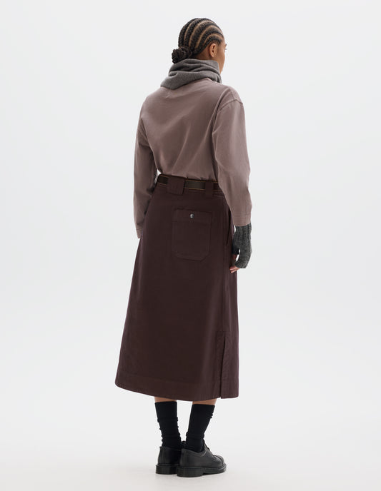Full body image, back view of model wearing dark brown A line skirt with side splits, belt loop detail on back and back patch pocket with reinforced stitch detailing. Styled with lilac long sleeve top tucked in, black leather belt, grey scarf and matching fingerless gloves. With black socks and black leather shoes.