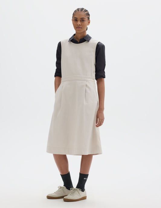 Full body image, front view of model wearing an off white cotton sleeveless pinafore style dress with side button fastening, a waistband with two pleats through the skirt part and slanted pockets. Styled with a black shirt, sleeves rolled, black socks and pale beige trainers.