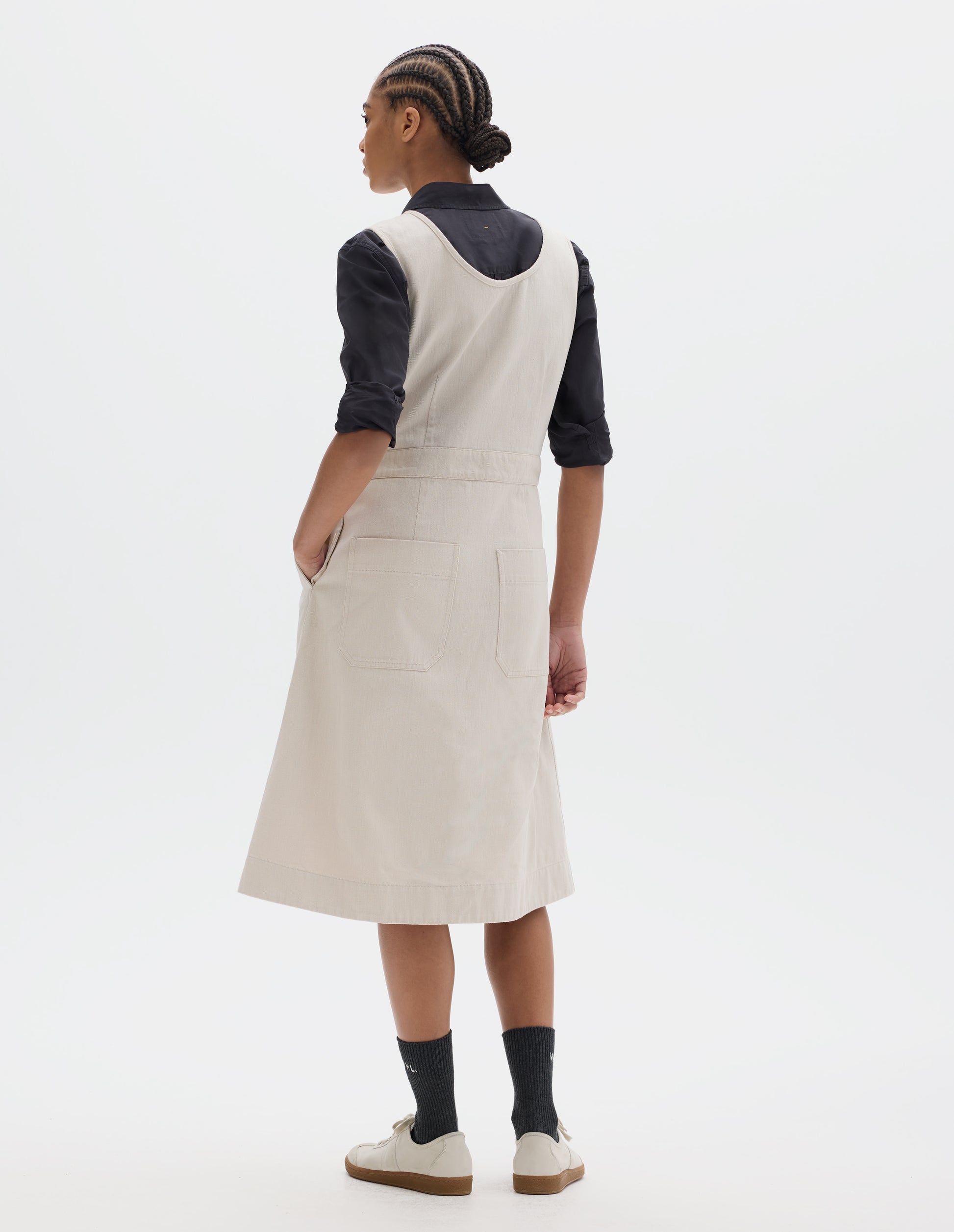 Full body image, back view of model wearing an off white cotton sleeveless pinafore style dress with a waistband, back darts through the skirt and two patch pockets. Styled with a black shirt, sleeves rolled, black socks and pale beige trainers.