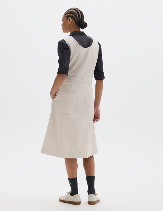 Full body image, back view of model wearing an off white cotton sleeveless pinafore style dress with a waistband, back darts through the skirt and two patch pockets. Styled with a black shirt, sleeves rolled, black socks and pale beige trainers.
