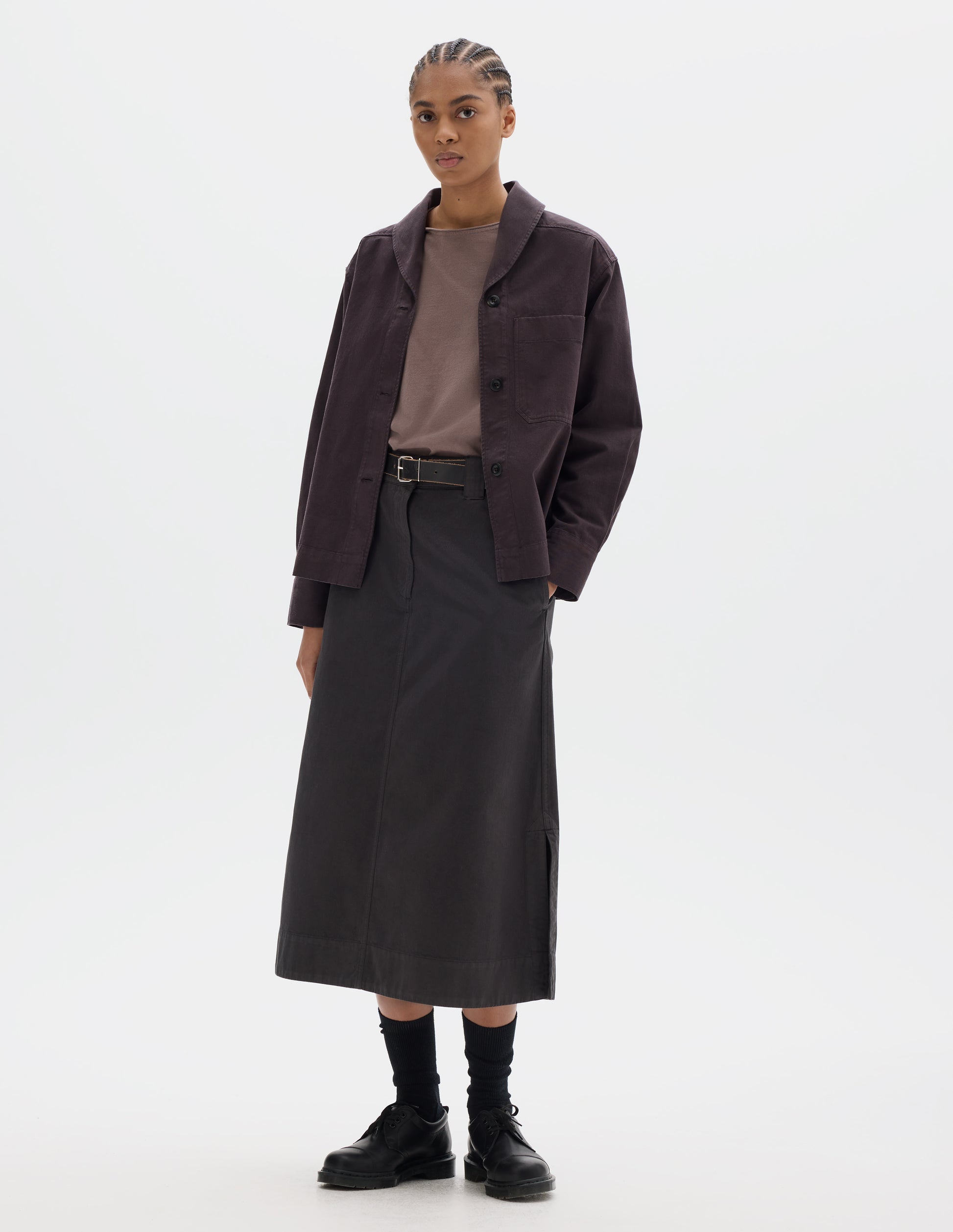 Full body image, front view of model wearing a dark brown cotton shawl collar overshirt with a front button placket, dark buttons, a forward-positioned shoulder seam and a front patch pocket. Styled with a grey pink jersey, dark brown cotton skirt, black leather belt and shoes and black socks.