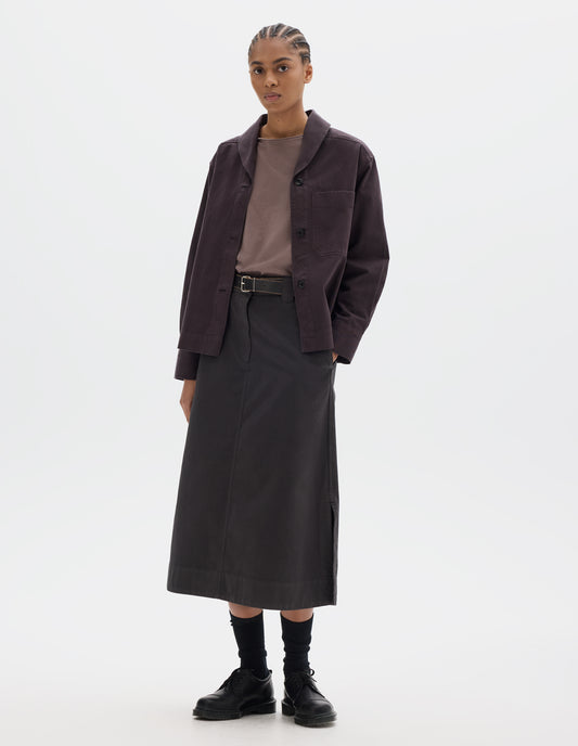 Full body image, front view of model wearing a dark brown cotton shawl collar overshirt with a front button placket, dark buttons, a forward-positioned shoulder seam and a front patch pocket. Styled with a grey pink jersey, dark brown cotton skirt, black leather belt and shoes and black socks.