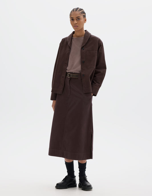 Full body image, front view of model wearing a dark brown cotton shawl collar overshirt with a front button placket, dark buttons, a forward-positioned shoulder seam and a front patch pocket. Styled with a grey pink jersey, dark brown cotton skirt, black leather belt and shoes and black socks.