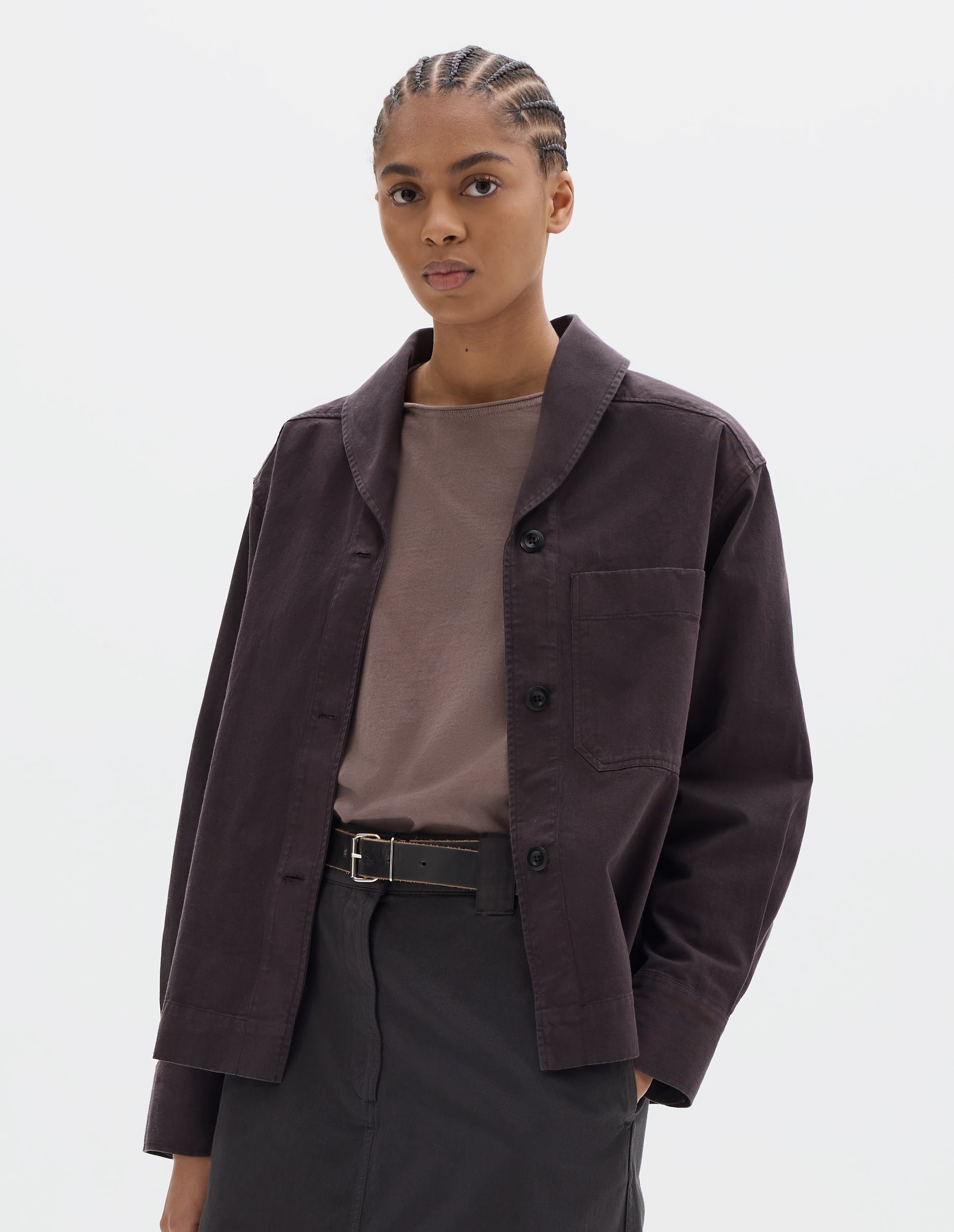Cropped body image, front view of model wearing a dark brown cotton shawl collar overshirt with a front button placket, dark buttons, a forward-positioned shoulder seam and a front patch pocket. Styled with a grey pink jersey, dark brown cotton skirt and black leather belt.