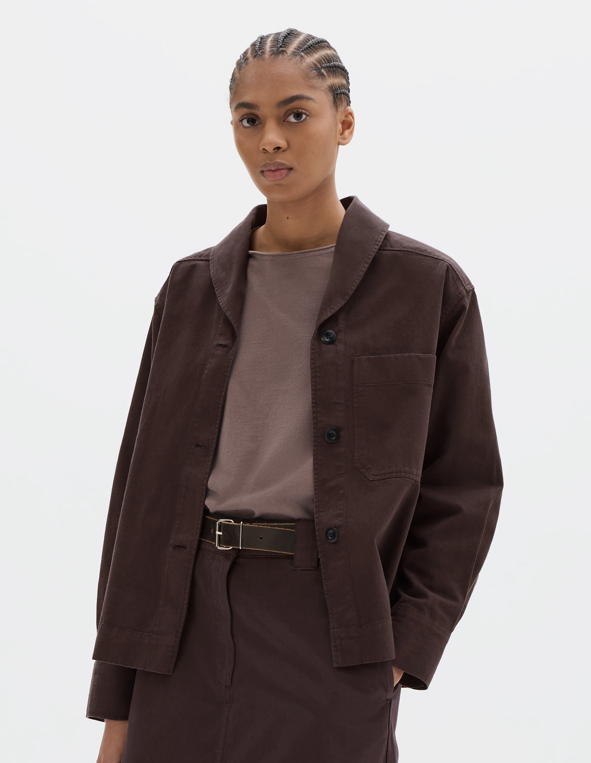 Cropped body image, front view of model wearing a dark brown cotton shawl collar overshirt with a front button placket, dark buttons, a forward-positioned shoulder seam and a front patch pocket. Styled with a grey pink jersey, dark brown cotton skirt and black leather belt.