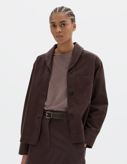 Cropped body image, front view of model wearing a dark brown cotton shawl collar overshirt with a front button placket, dark buttons, a forward-positioned shoulder seam and a front patch pocket. Styled with a grey pink jersey, dark brown cotton skirt and black leather belt.