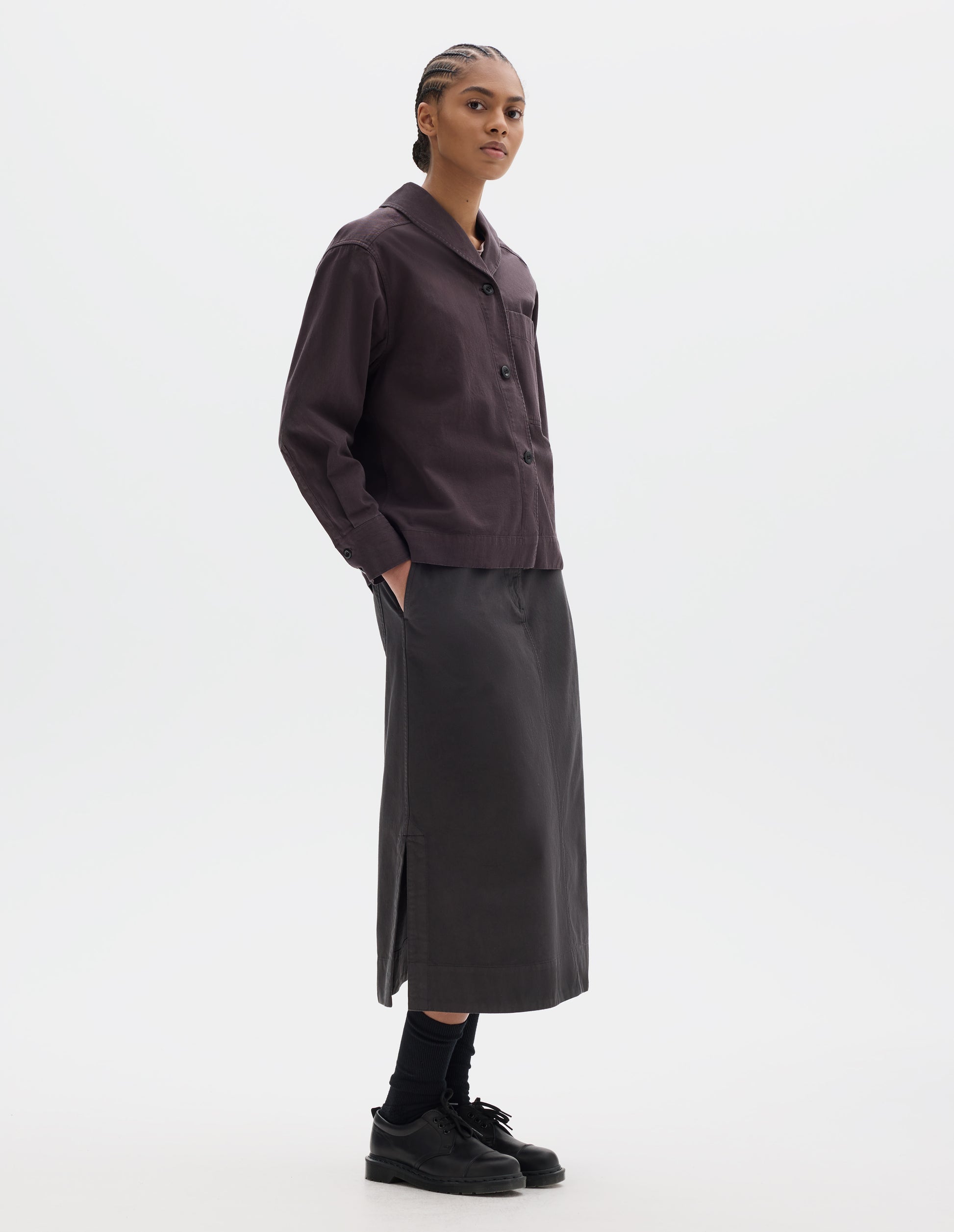Full body image, front side view of model wearing a dark brown cotton shawl collar overshirt with a front button placket, a forward-positioned shoulder seam and shirt cuffs with double button fastening. Styled with a dark brown cotton skirt, black leather belt and shoes and black socks.