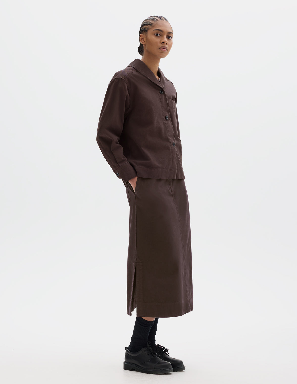 Full body image, front side view of model wearing a dark brown cotton shawl collar overshirt with a front button placket, a forward-positioned shoulder seam and shirt cuffs with double button fastening. Styled with a dark brown cotton skirt, black leather belt and shoes and black socks.