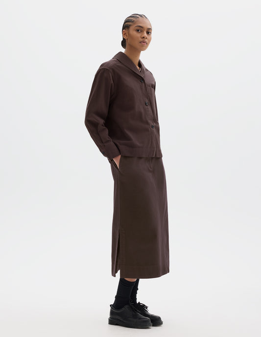 Full body image, front side view of model wearing a dark brown cotton shawl collar overshirt with a front button placket, a forward-positioned shoulder seam and shirt cuffs with double button fastening. Styled with a dark brown cotton skirt, black leather belt and shoes and black socks.