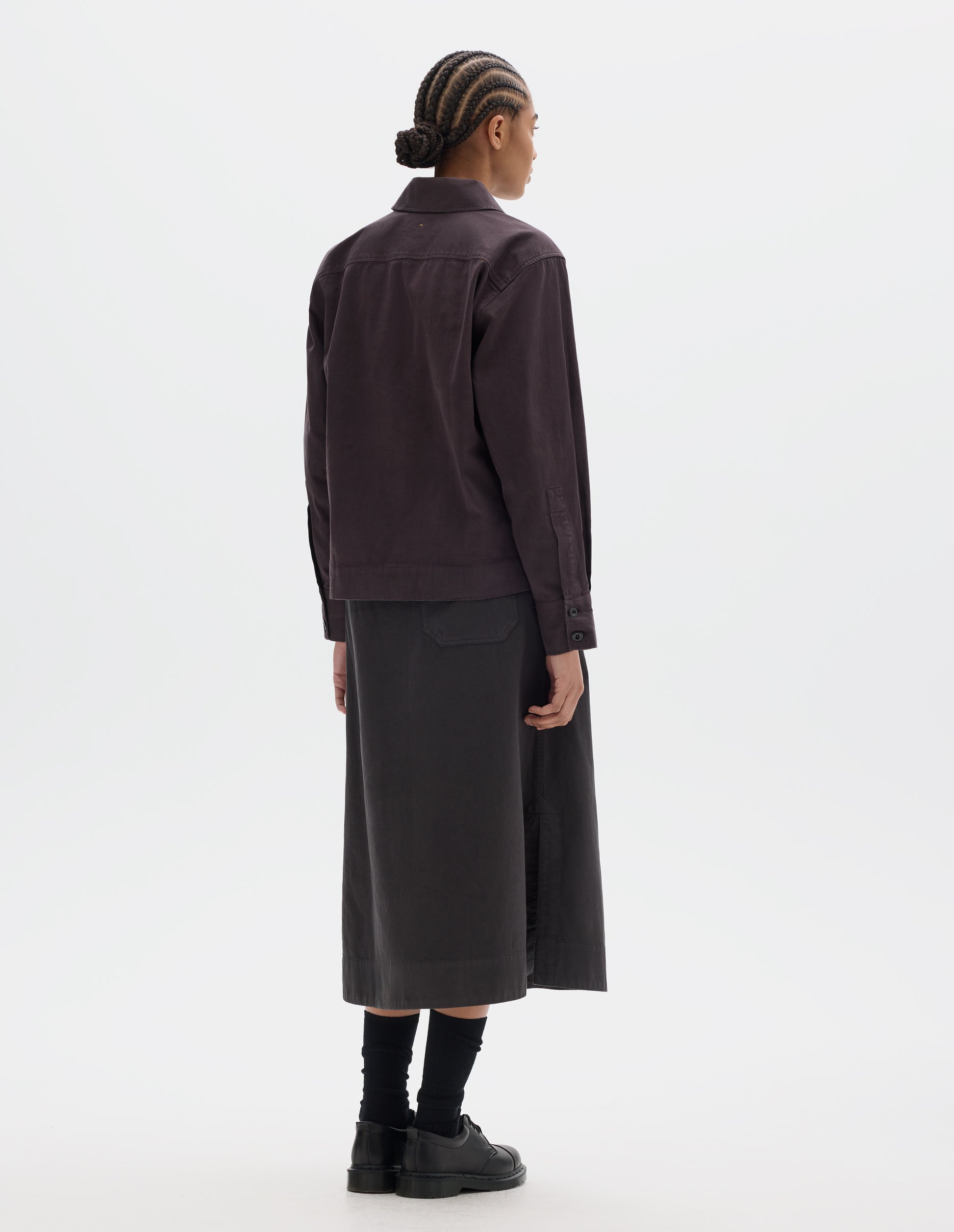 Full body image, back view of model wearing a dark brown cotton shawl collar overshirt with a forward-positioned shoulder seam, back yoke detailing and cuffs with double button fastening. Styled with a dark brown cotton skirt, black leather belt and shoes and black socks.