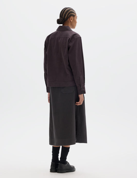 Full body image, back view of model wearing a dark brown cotton shawl collar overshirt with a forward-positioned shoulder seam, back yoke detailing and cuffs with double button fastening. Styled with a dark brown cotton skirt, black leather belt and shoes and black socks.