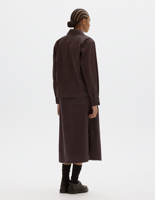 Full body image, back view of model wearing a dark brown cotton shawl collar overshirt with a forward-positioned shoulder seam, back yoke detailing and cuffs with double button fastening. Styled with a dark brown cotton skirt, black leather belt and shoes and black socks.