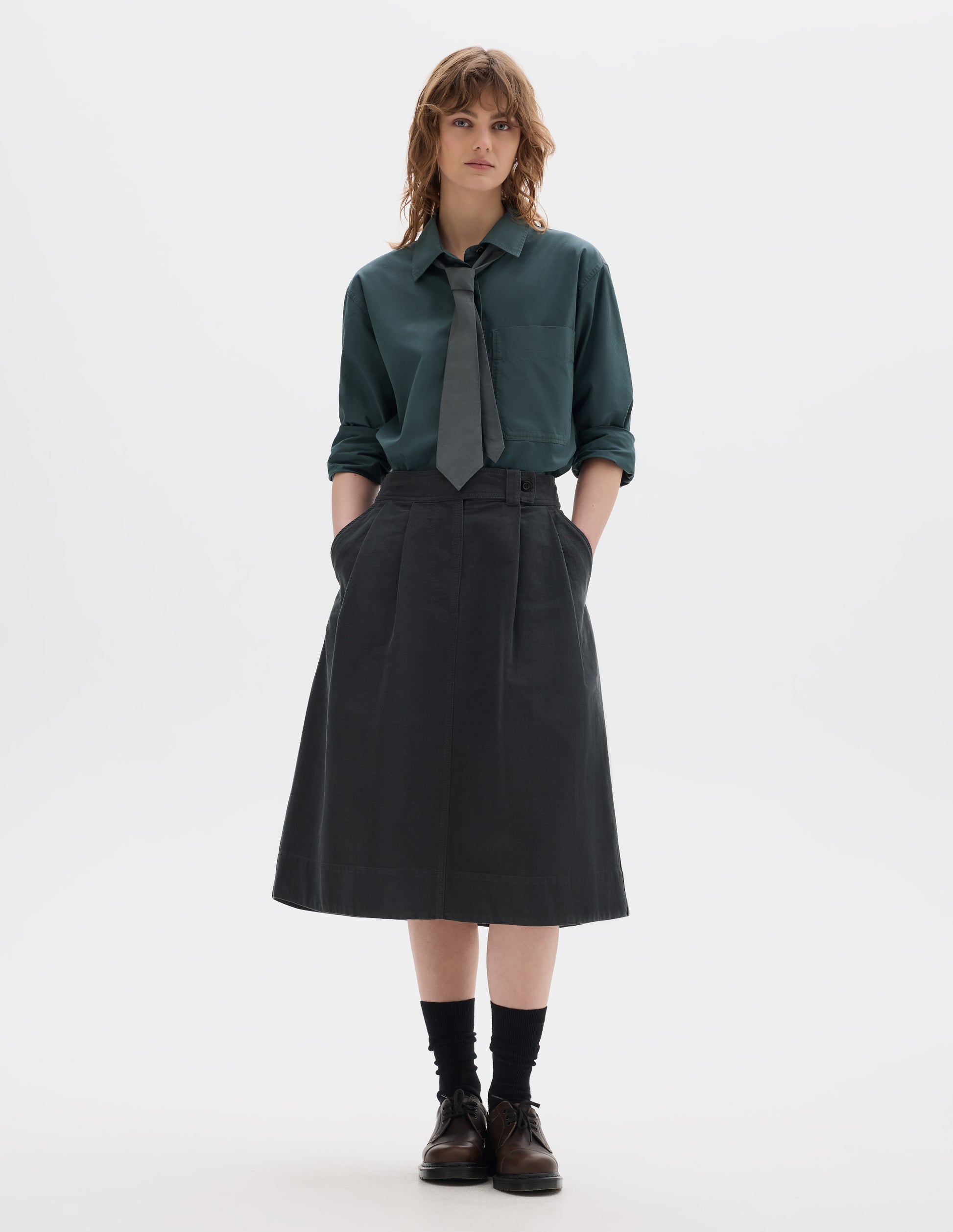 Full body image, front view of model wearing a blue green shirt with a front patch pocket, sleeves rolled up, dark green greyish pleated a line skirt, mid-calf length, constructed with an extended waistband with a loop and button fastening and slanted front pockets. Styled with a greyish green tie, black socks and brown leather shoes.