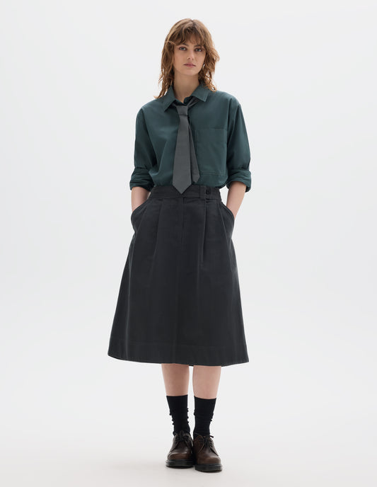 Full body image, front view of model wearing a blue green shirt with a front patch pocket, sleeves rolled up, dark green greyish pleated a line skirt, mid-calf length, constructed with an extended waistband with a loop and button fastening and slanted front pockets. Styled with a greyish green tie, black socks and brown leather shoes.