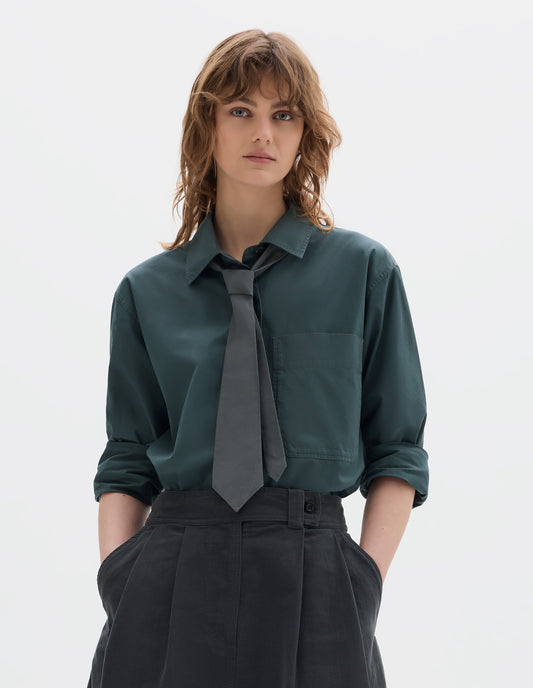 Cropped body image, front view of model wearing a blue green shirt with a front patch pocket, sleeves rolled up, styled with a greyish green tie and a dark green greyish pleated a line skirt.
