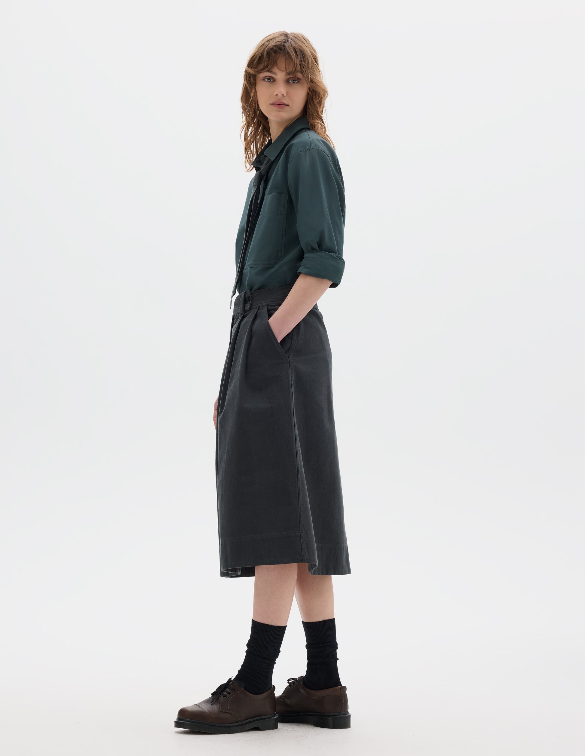 Full body image, front side view of model wearing a blue green shirt with a front patch pocket, sleeves rolled up, dark green greyish pleated a line skirt, mid-calf length, constructed with an extended waistband with a loop and button fastening and slanted front pockets. Styled with a greyish green tie, black socks and brown leather shoes.