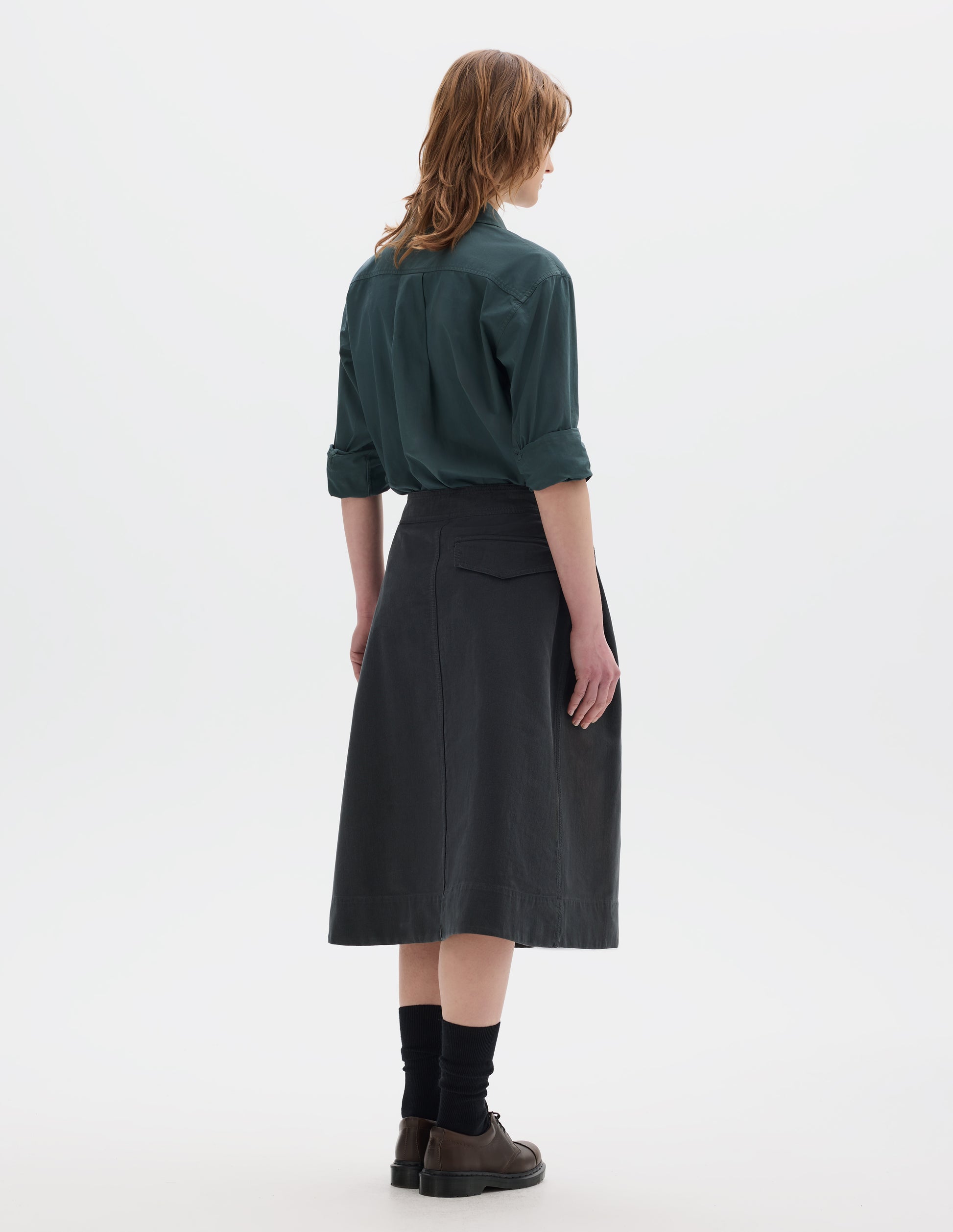 Full body image, back view of model wearing a blue green shirt, sleeves rolled up, dark green greyish pleated a line skirt, mid-calf length with a concealed button-fastening back flap pocket. Styled with black socks and brown leather shoes.