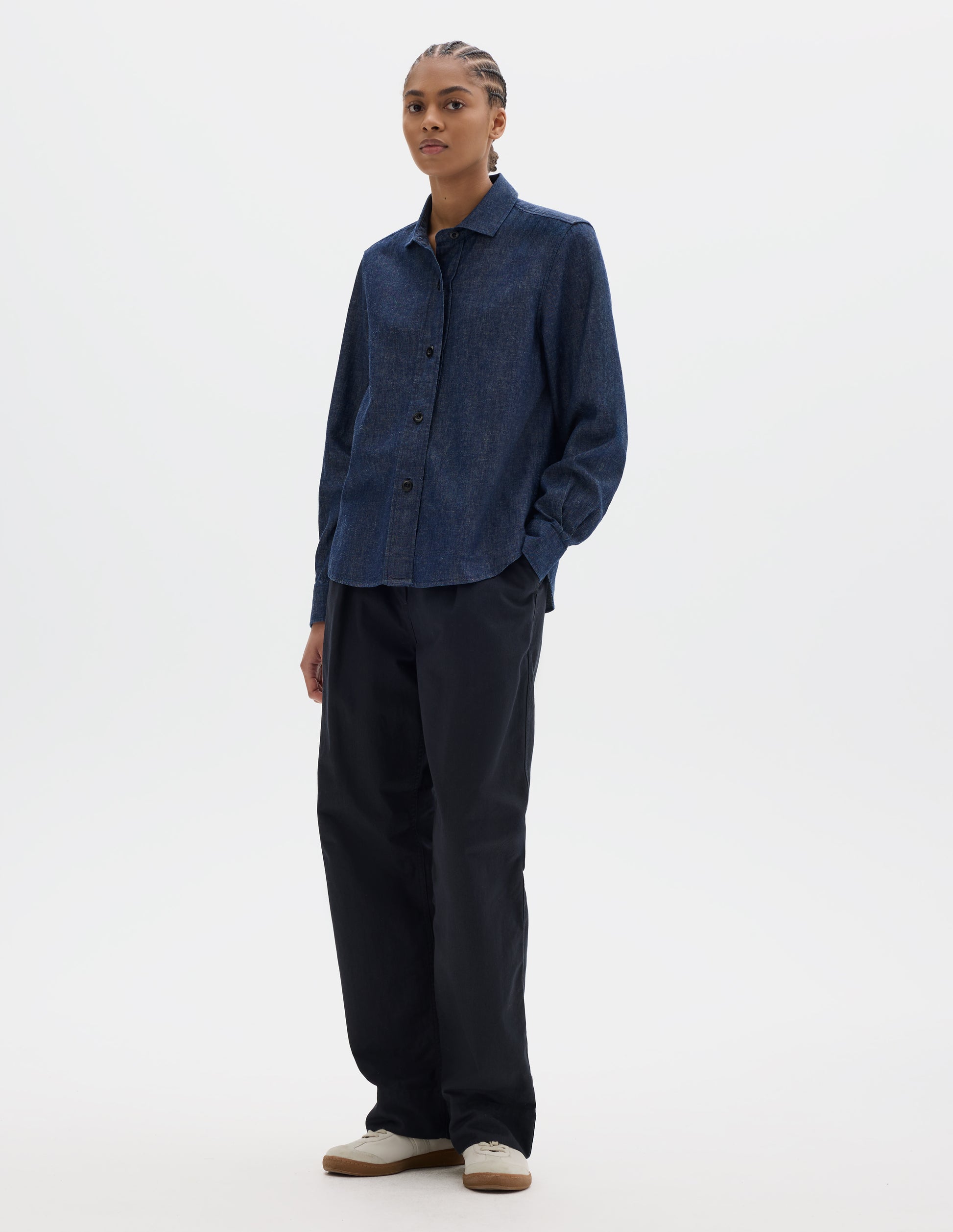 Full body image, front view of model wearing a dark blue shirt with a stitch-through raised placket, curved hem and a button-through cuff gauntlet. Styled with dark blue trousers and pale beige trainers.