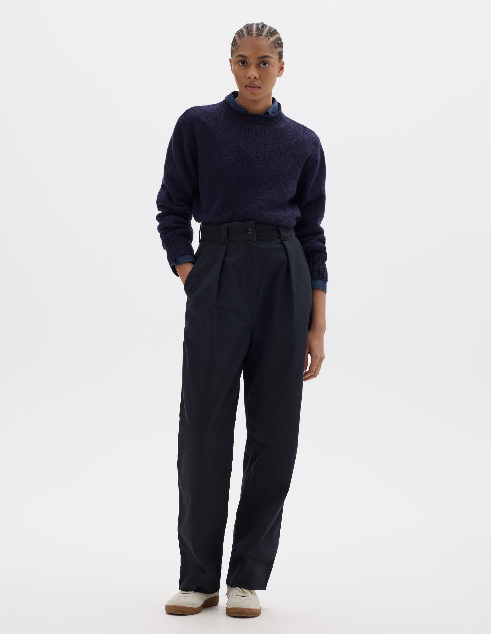 Fully body image, front view of model wearing dark blue cotton trousers, with front pleats, a dark button with belt loops and slanted side pockets. Styled with navy shirt, navy jumper and white shoes.