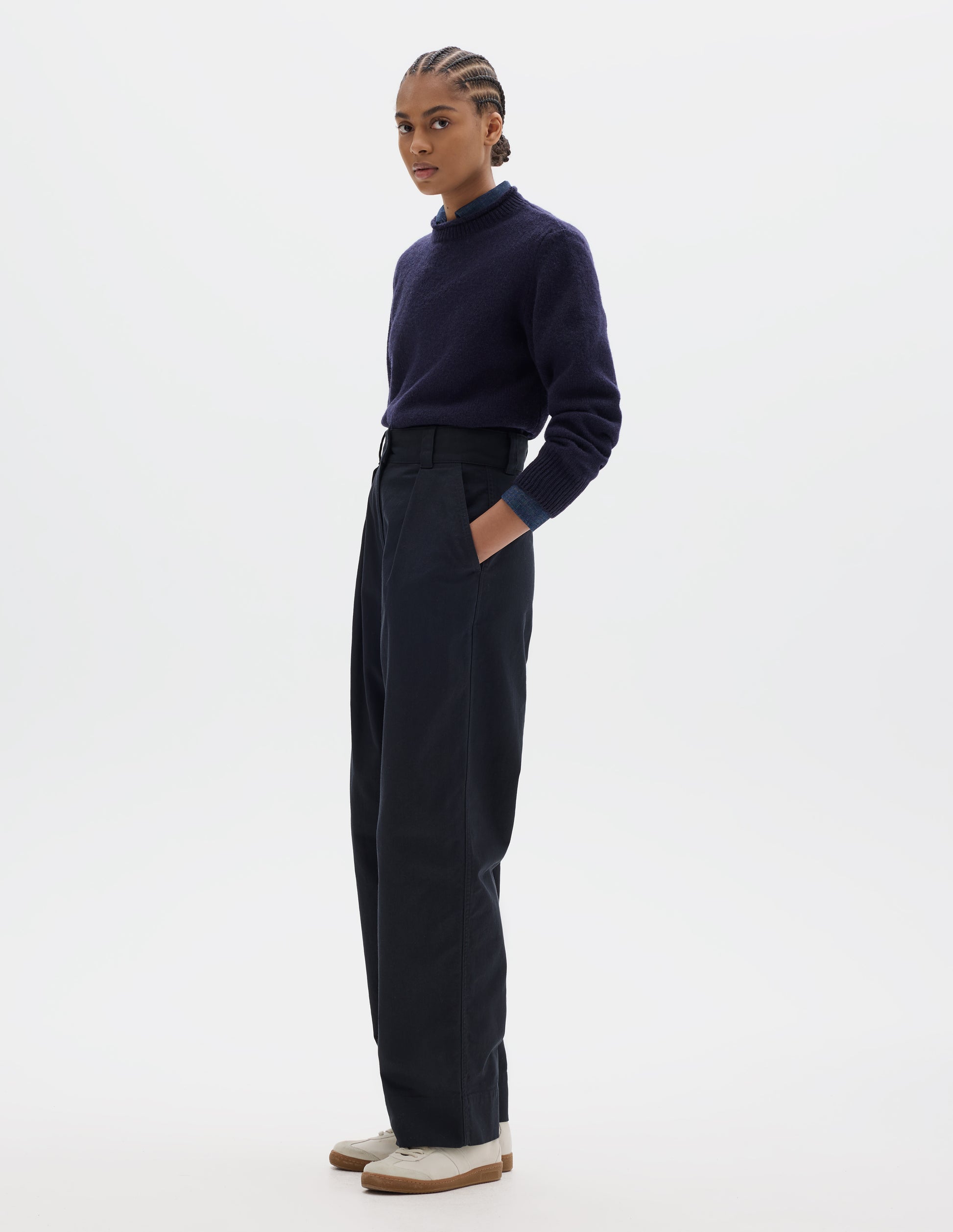 Fully body image, side view of model wearing dark blue cotton trousers, with front pleats, a dark button with belt loops and slanted side pockets. Styled with navy shirt, navy jumper and white shoes.