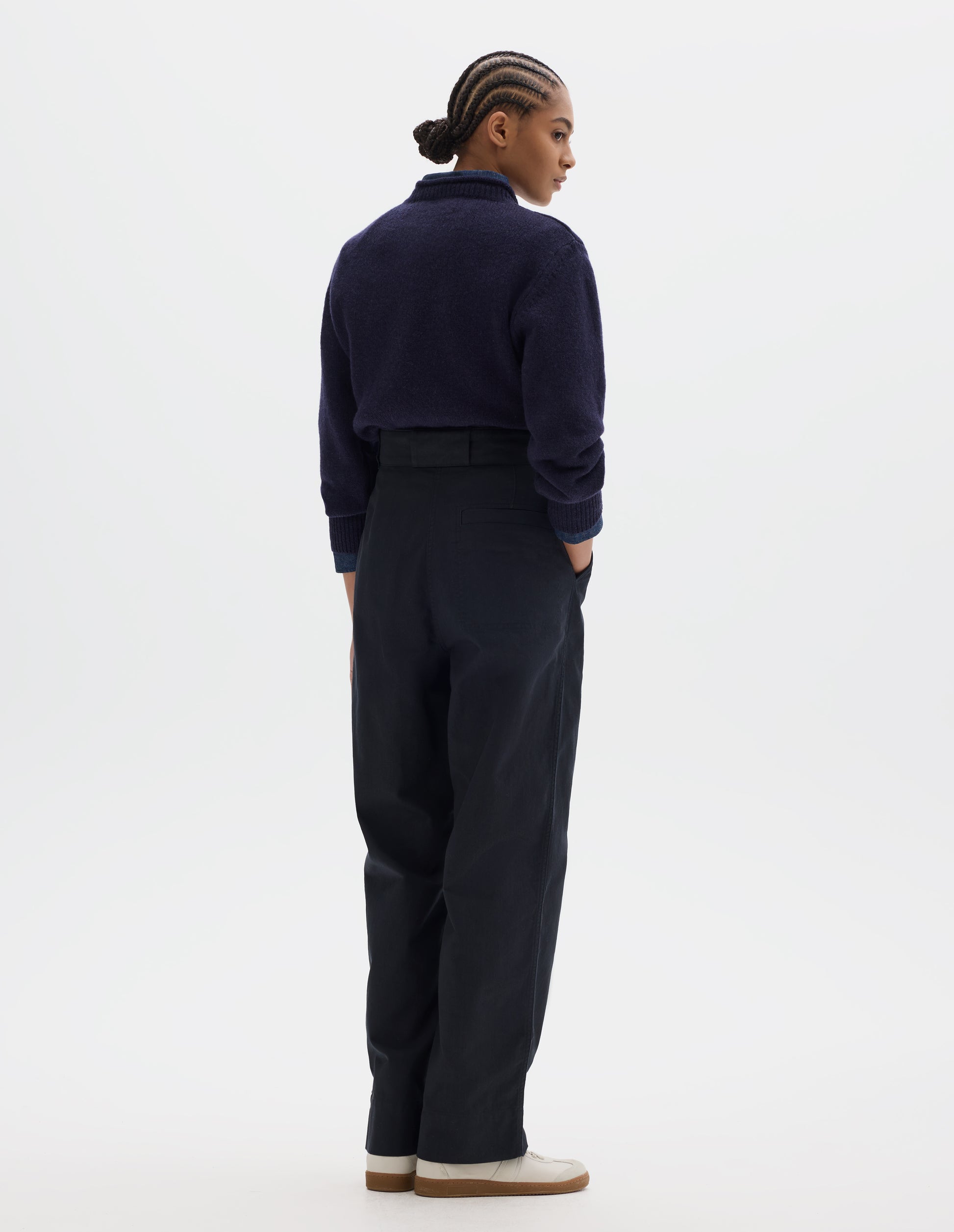 Fully body image, back view of model wearing dark blue cotton trousers, with belt loops and a single welt pocket. Styled with navy shirt, navy jumper and white shoes. 