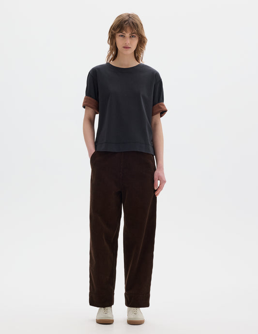 Full body image, front view of model wearing a brown cropped and flat fronted corduroy trousers with deep hem and slanted side pockets. Styled with a short sleeve T shirt in black with a contrast brown colour on the reverse of the sleeve and off white suede trainers.