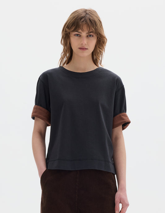 Cropped body image, front view model wearing a short sleeve T shirt in black constructed with a wide neck, deep faced cuffs in with a contrast  brown colour on the reverse, and flatlock stitching on the hem and armholes. Styled with brown corduroy trousers.