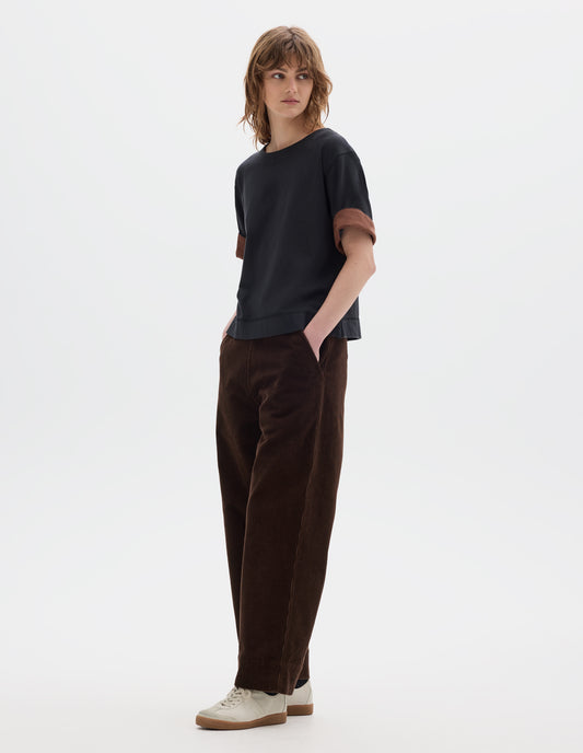 Full body image, front view of model wearing a short sleeve T shirt in black constructed with a wide neck, deep faced cuffs in with a contrast brown colour on the reverse, and flatlock stitching on the hem and armholes. Styled with brown corduroy trousers and white suede trainers.