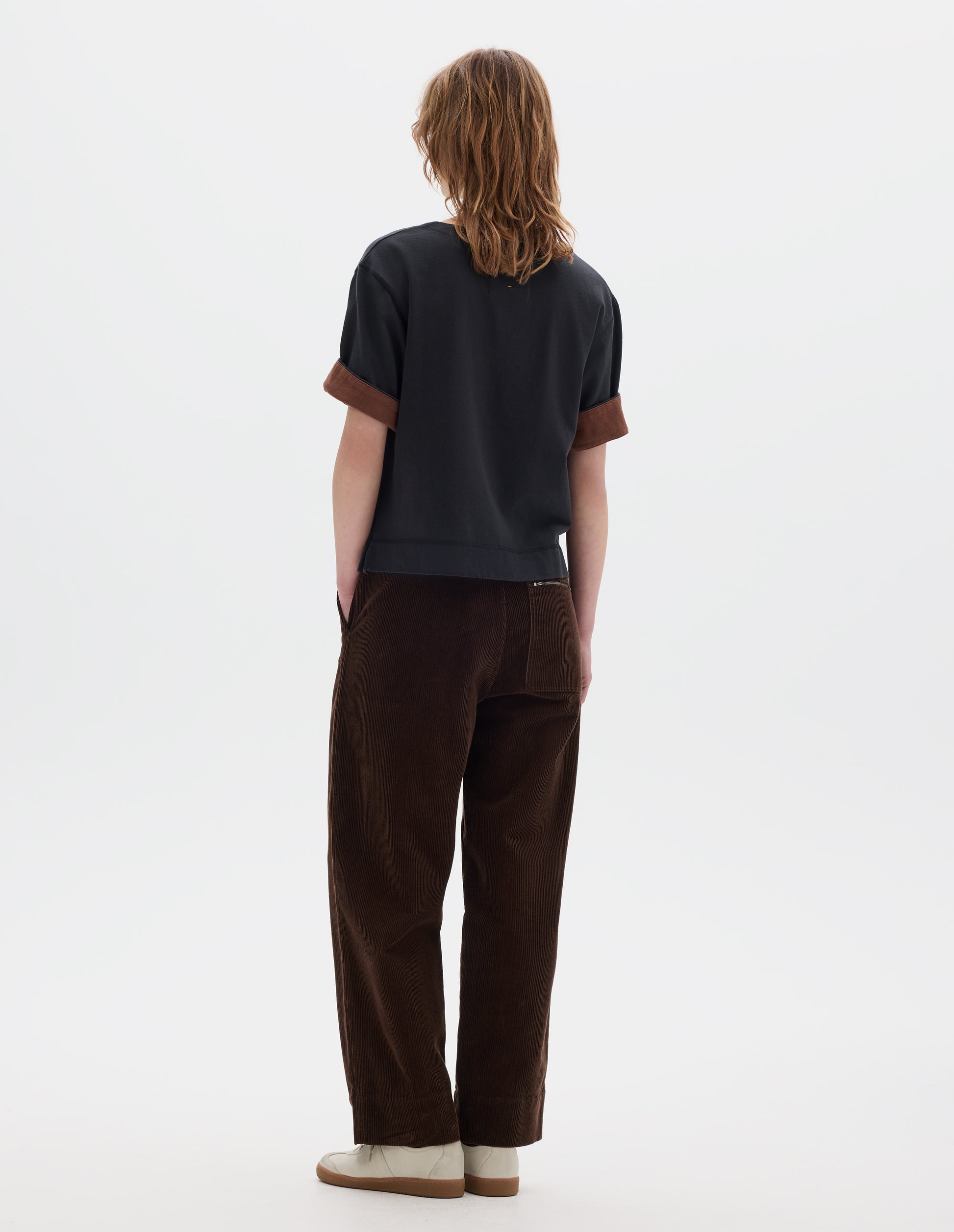 Full body image, back view of model wearing a short sleeve T shirt in black constructed with a wide neck, deep faced cuffs in with a contrast brown colour on the reverse, and flatlock stitching on the hem and armholes. Styled with brown corduroy trousers and white suede trainers.