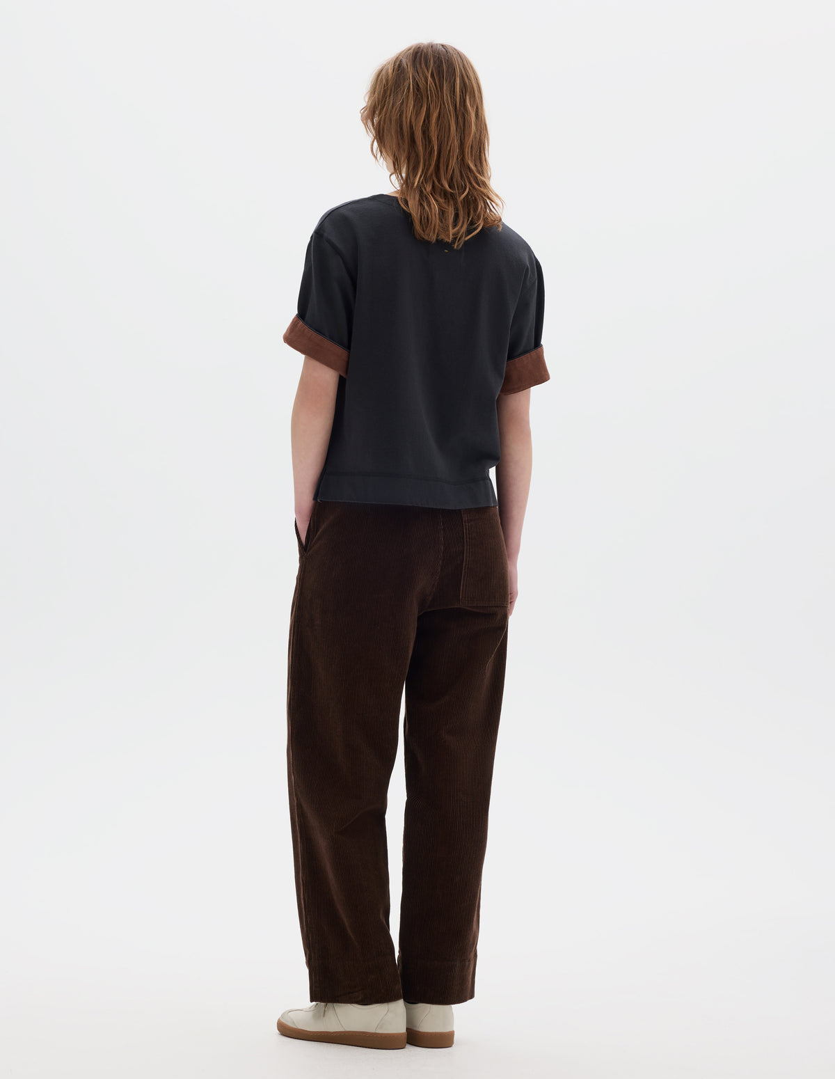 Full body image, back view of model wearing a brown cropped corduroy trousers with deep hem, slanted side pockets and a back patch pocket. Styled with a short sleeve T shirt in black with a contrast brown colour on the reverse of the sleeve and off white suede trainers.