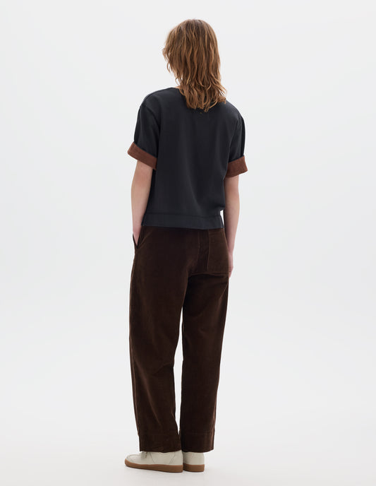 Full body image, back view of model wearing a brown cropped corduroy trousers with deep hem, slanted side pockets and a back patch pocket. Styled with a short sleeve T shirt in black with a contrast brown colour on the reverse of the sleeve and off white suede trainers.
