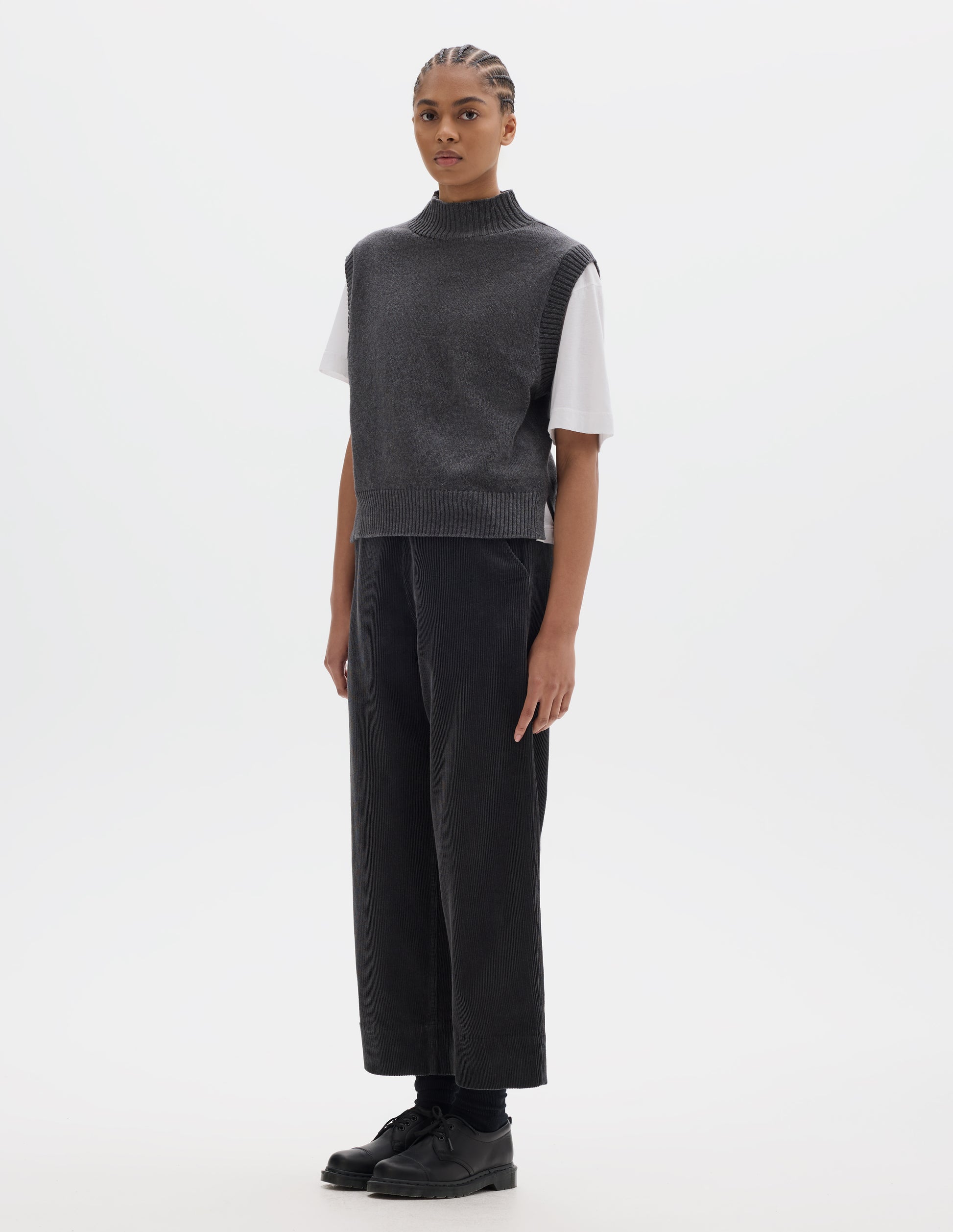 Full body image, front view of model wearing a grey wool cotton slipover with a high rib neck, rib armholes and hem and split stepped hem. Styled over a white t shirt and dark grey corduroy trousers, black socks and leather shoes.