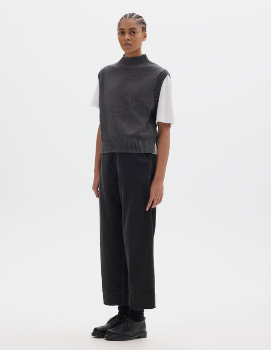 Full body image, front view of model wearing a grey wool cotton slipover with a high rib neck, rib armholes and hem and split stepped hem. Styled over a white t shirt and dark grey corduroy trousers, black socks and leather shoes.