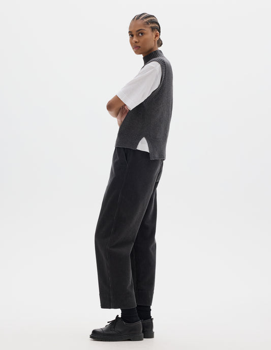 Full body image, front side view of model wearing a grey wool cotton slipover with a high rib neck, rib armholes and hem and split stepped hem. Styled over a white t shirt and dark grey corduroy trousers, black socks and leather shoes.