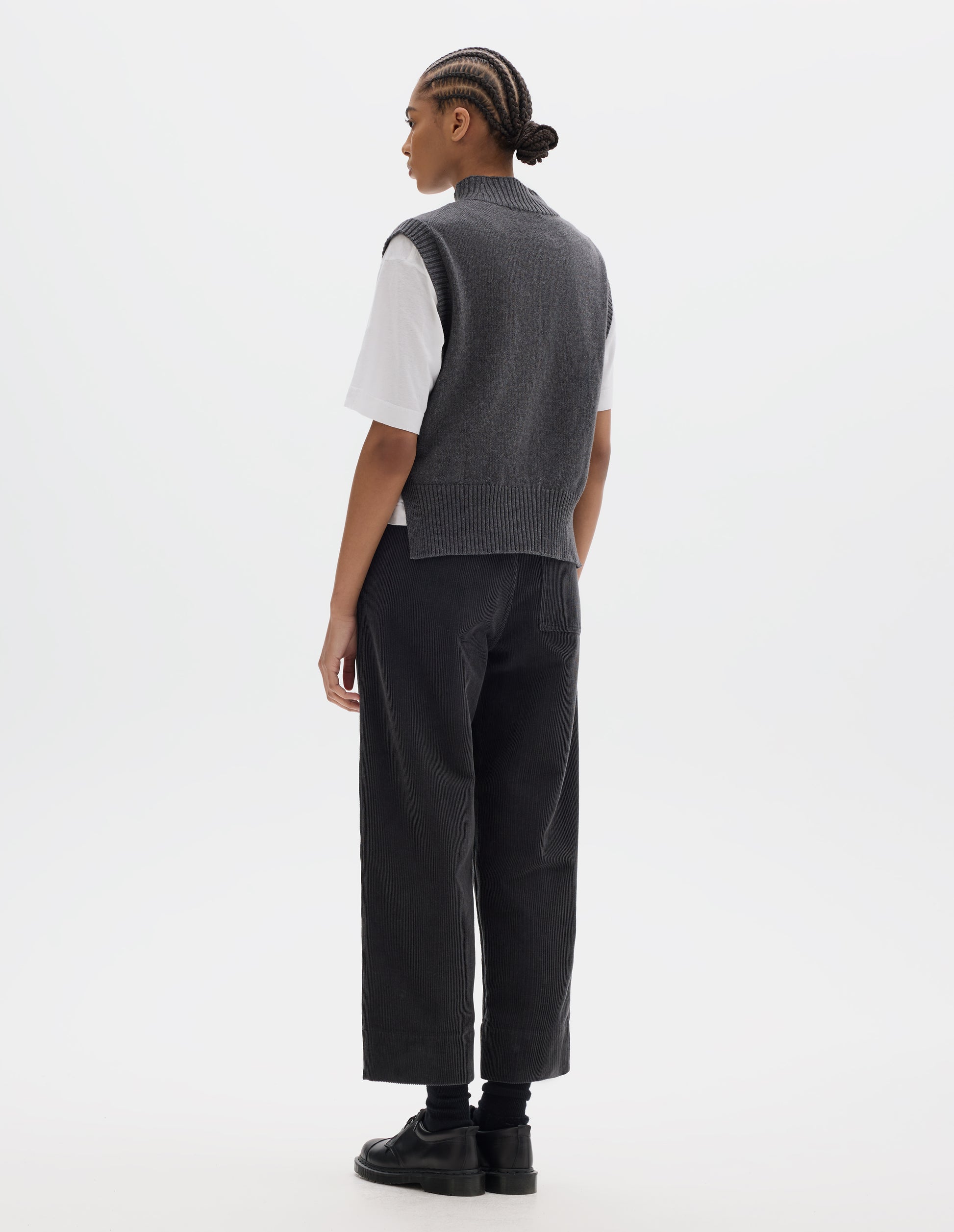 Full body image, back view of model wearing a grey wool cotton slipover with a high rib neck, rib armholes and hem and split stepped hem. Styled over a white t shirt and dark grey corduroy trousers, black socks and leather shoes.