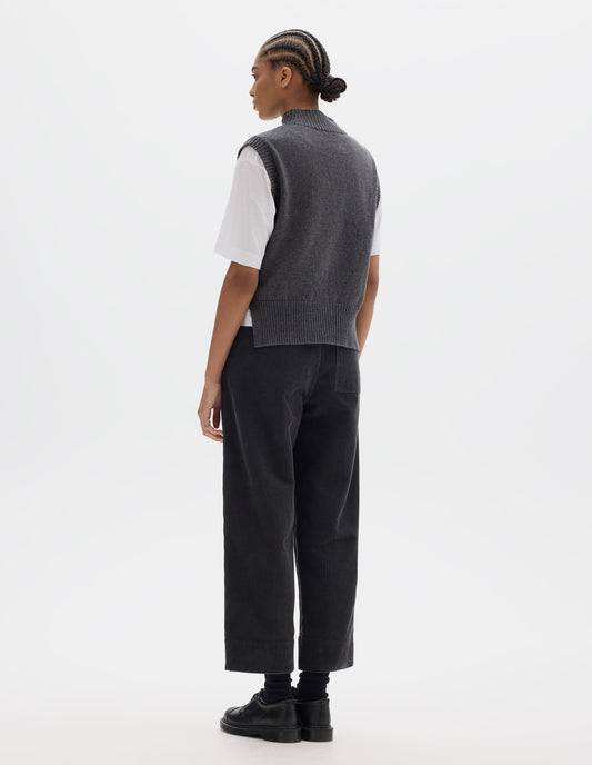 Full body image, back view of model wearing a grey wool cotton slipover with a high rib neck, rib armholes and hem and split stepped hem. Styled over a white t shirt and dark grey corduroy trousers, black socks and leather shoes.