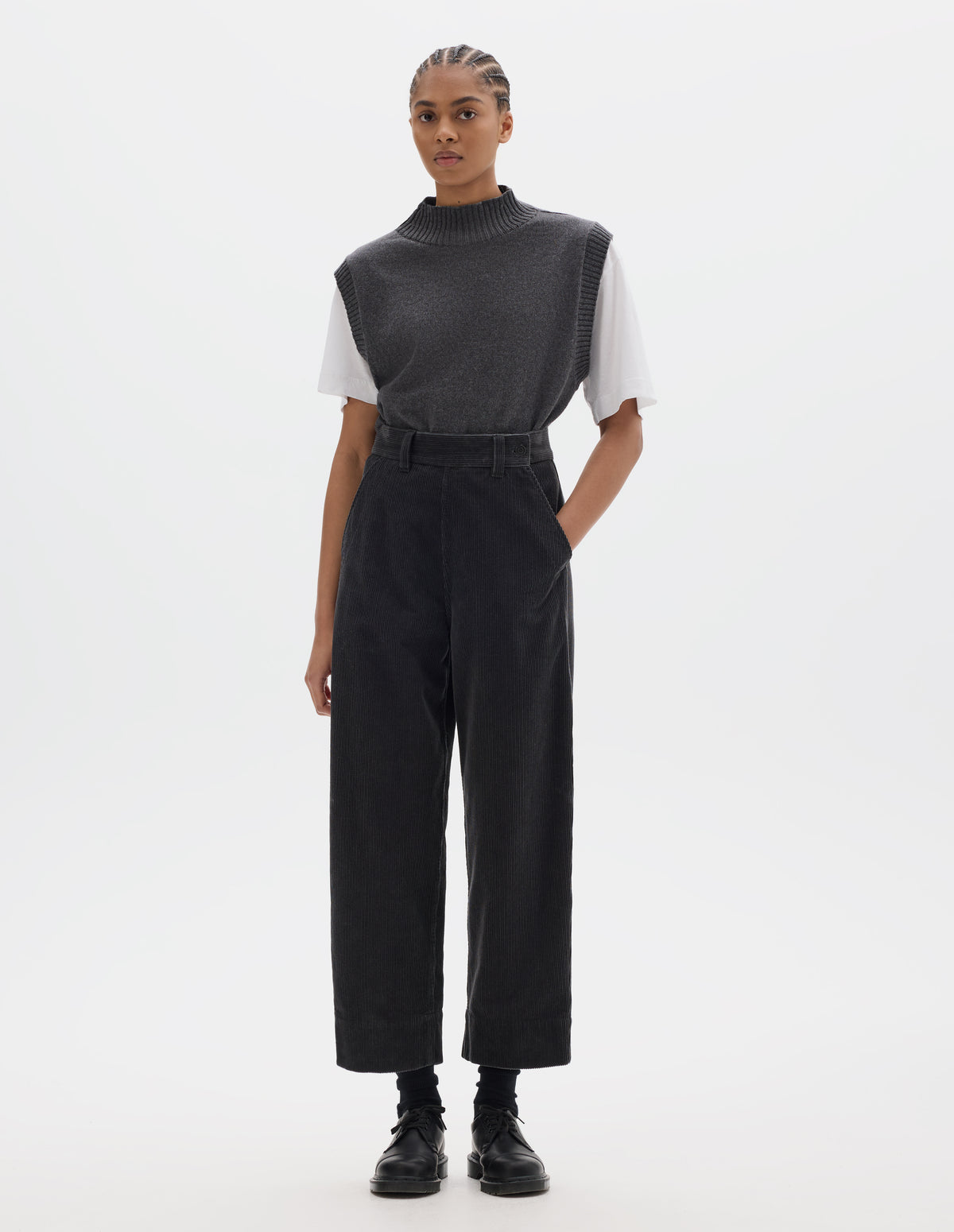 Full body image, front view of model wearing dark grey cotton corduroy trousers, with offset button fastening waistband, flat front side opening, slanted pocket and deep hem. Styled with white t-shirt and grey slipover tucked in, with black socks and black leather shoes.