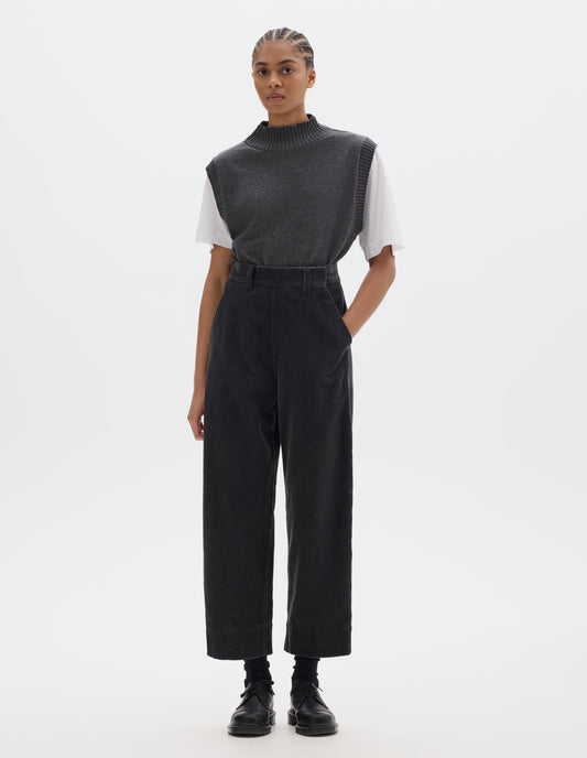 Full body image, front view of model wearing dark grey cotton corduroy trousers, with offset button fastening waistband, flat front side opening, slanted pocket and deep hem. Styled with white t-shirt and grey slipover tucked in, with black socks and black leather shoes.