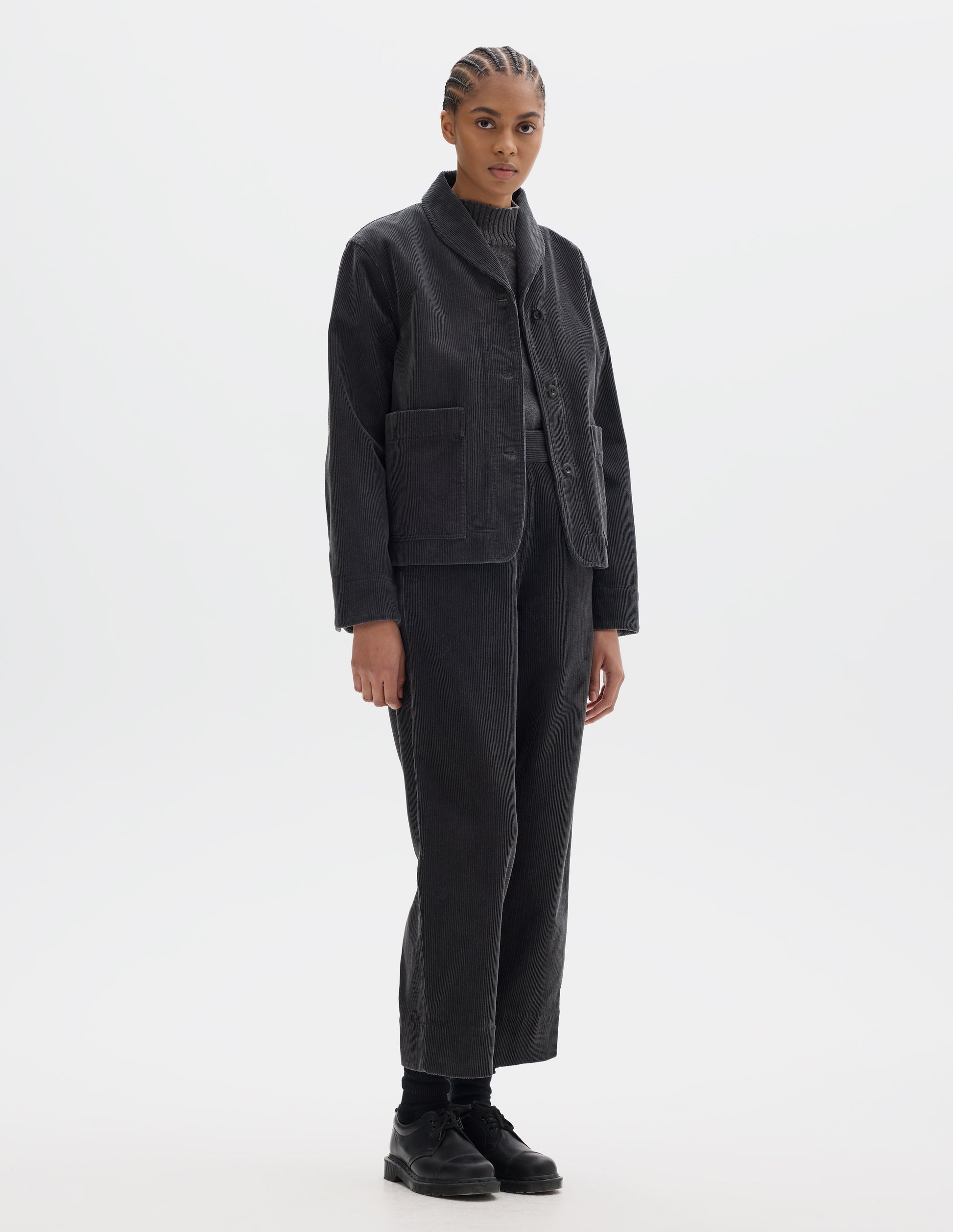 Full body image, front view of model wearing dark grey cotton corduroy jacket, with one piece collar, dropped shoulder, stitch-through placket, large patch pockets and corozo button closure and shirt cuffs. Styled over grey slipover and with matching dark grey corduroy trousers, with black socks and black leather shoes.