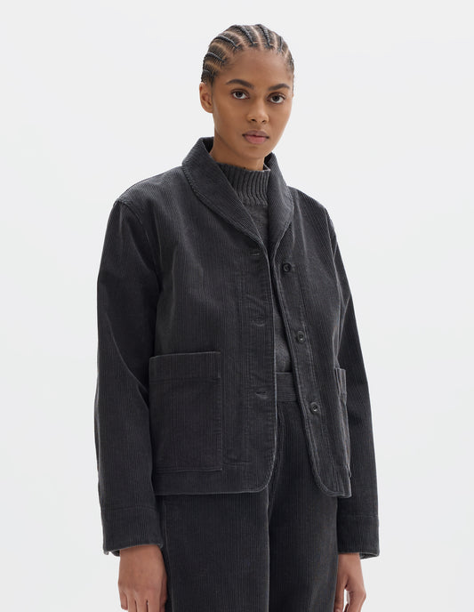 Cropped body image, front view of model wearing dark grey cotton corduroy jacket, with one piece collar, dropped shoulder, stitch-through placket, large patch pockets and corozo button closure and shirt cuffs. Styled over grey slipover and with matching dark grey corduroy trousers.