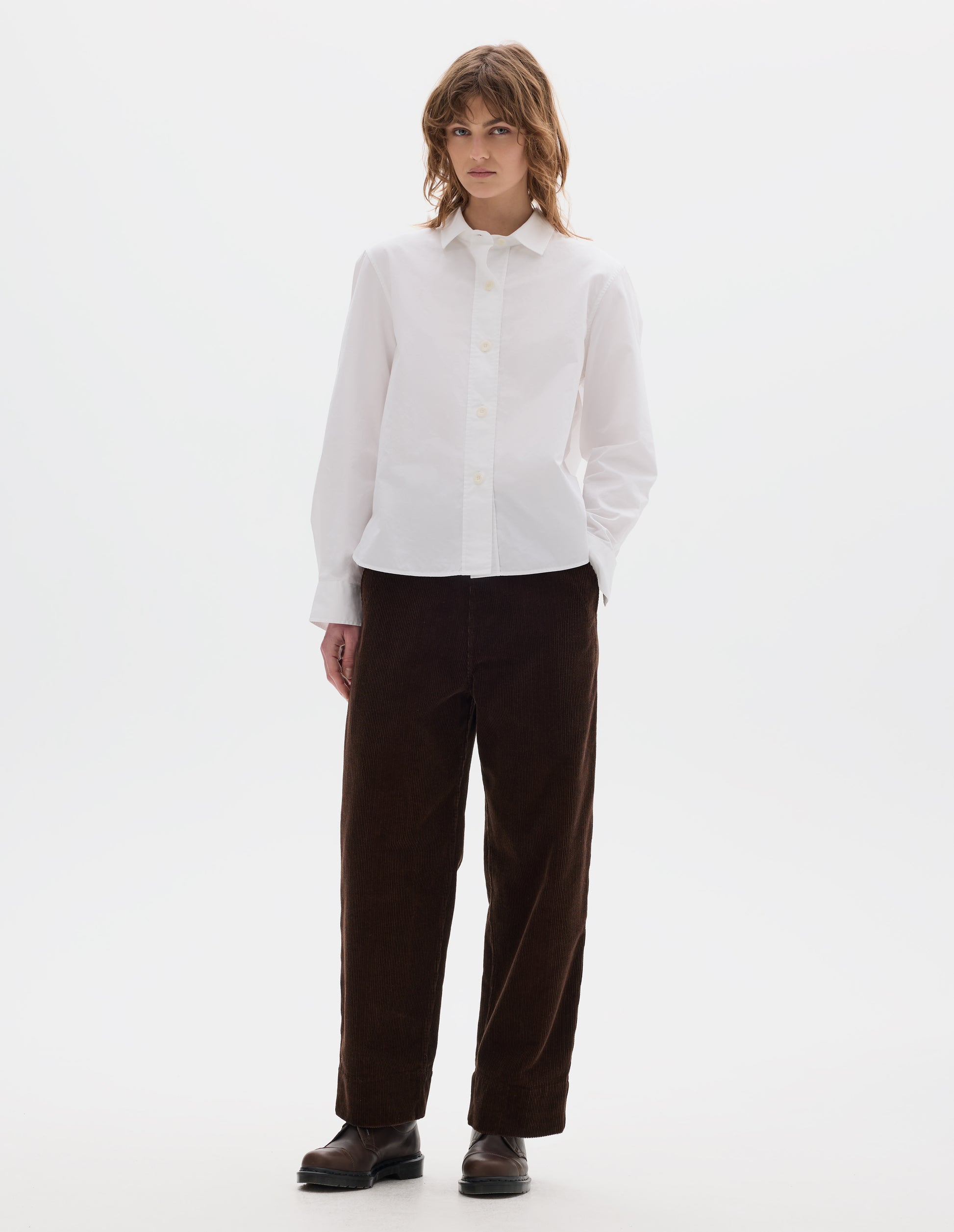 Full body image, front view of model wearing white regular cut shirt with stitch through raised placket, corozo buttons on placket and cuffs, curved hem. Styled with brown corduroy trousers and brown leather shoes.