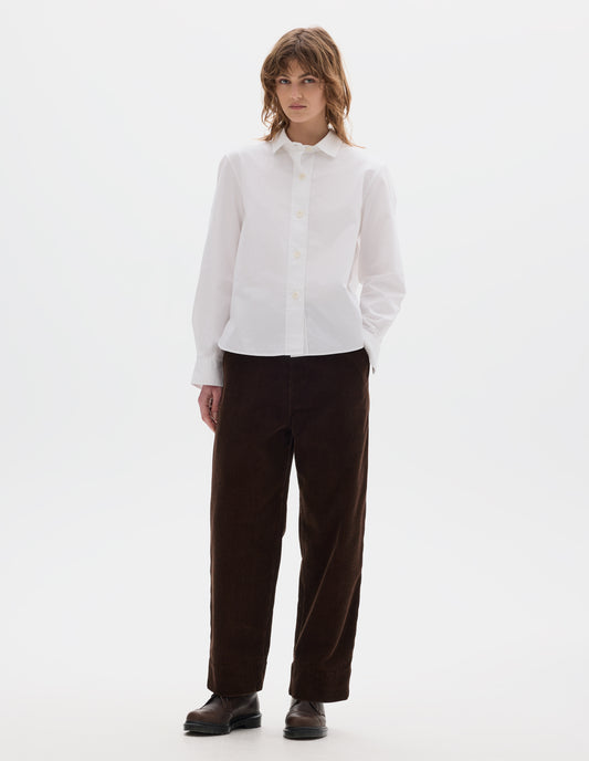 Full body image, front view of model wearing white regular cut shirt with stitch through raised placket, corozo buttons on placket and cuffs, curved hem. Styled with brown corduroy trousers and brown leather shoes.
