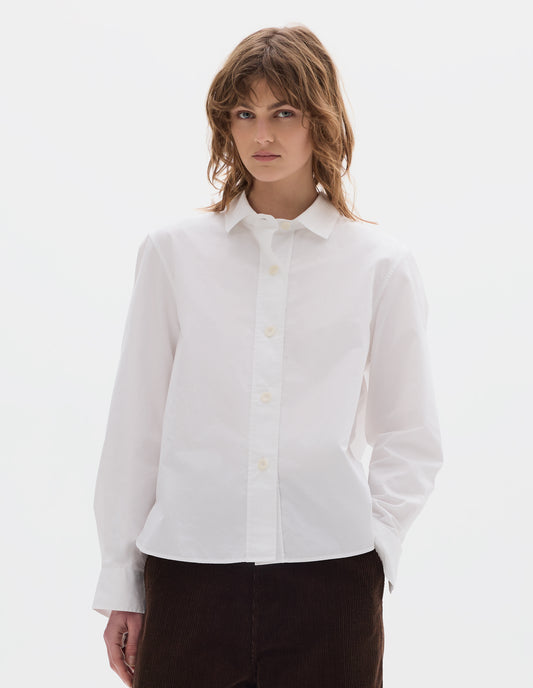 Cropped body image, front view of model wearing white regular cut shirt with stitch through raised placket, corozo buttons on placket and cuffs, curved hem. Styled with brown corduroy trousers.