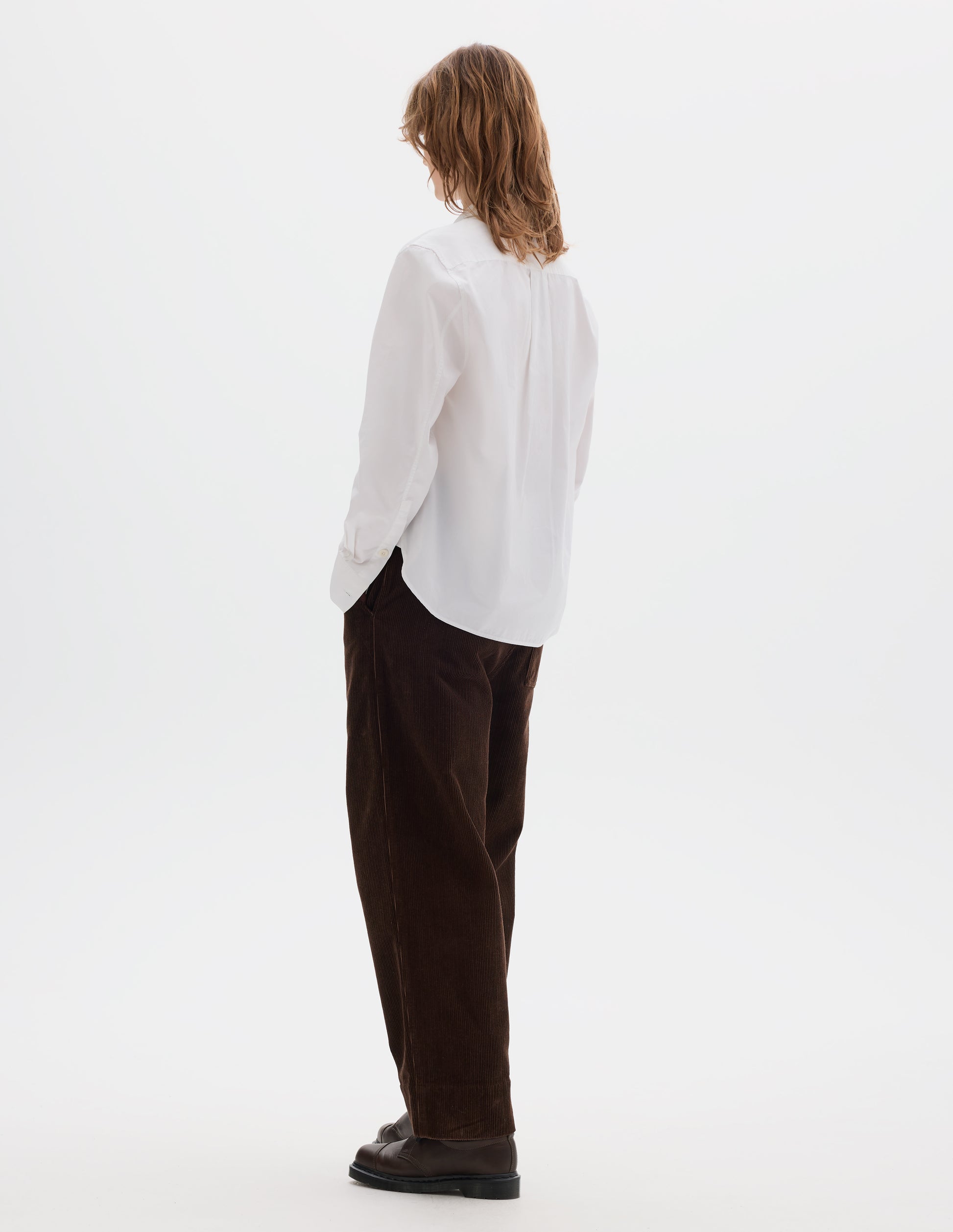 Full body image, back view of model wearing white regular cut shirt with box pleat on back yoke, corozo button cuffs and curved hem. Styled with brown corduroy trousers and brown leather shoes.