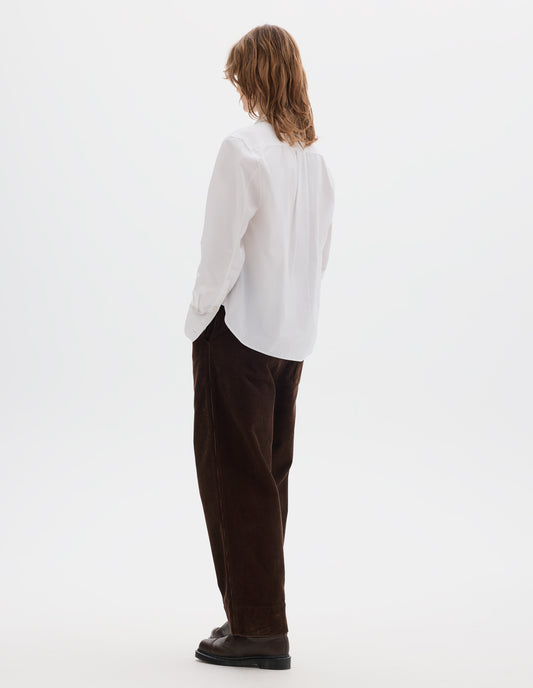 Full body image, back view of model wearing white regular cut shirt with box pleat on back yoke, corozo button cuffs and curved hem. Styled with brown corduroy trousers and brown leather shoes.