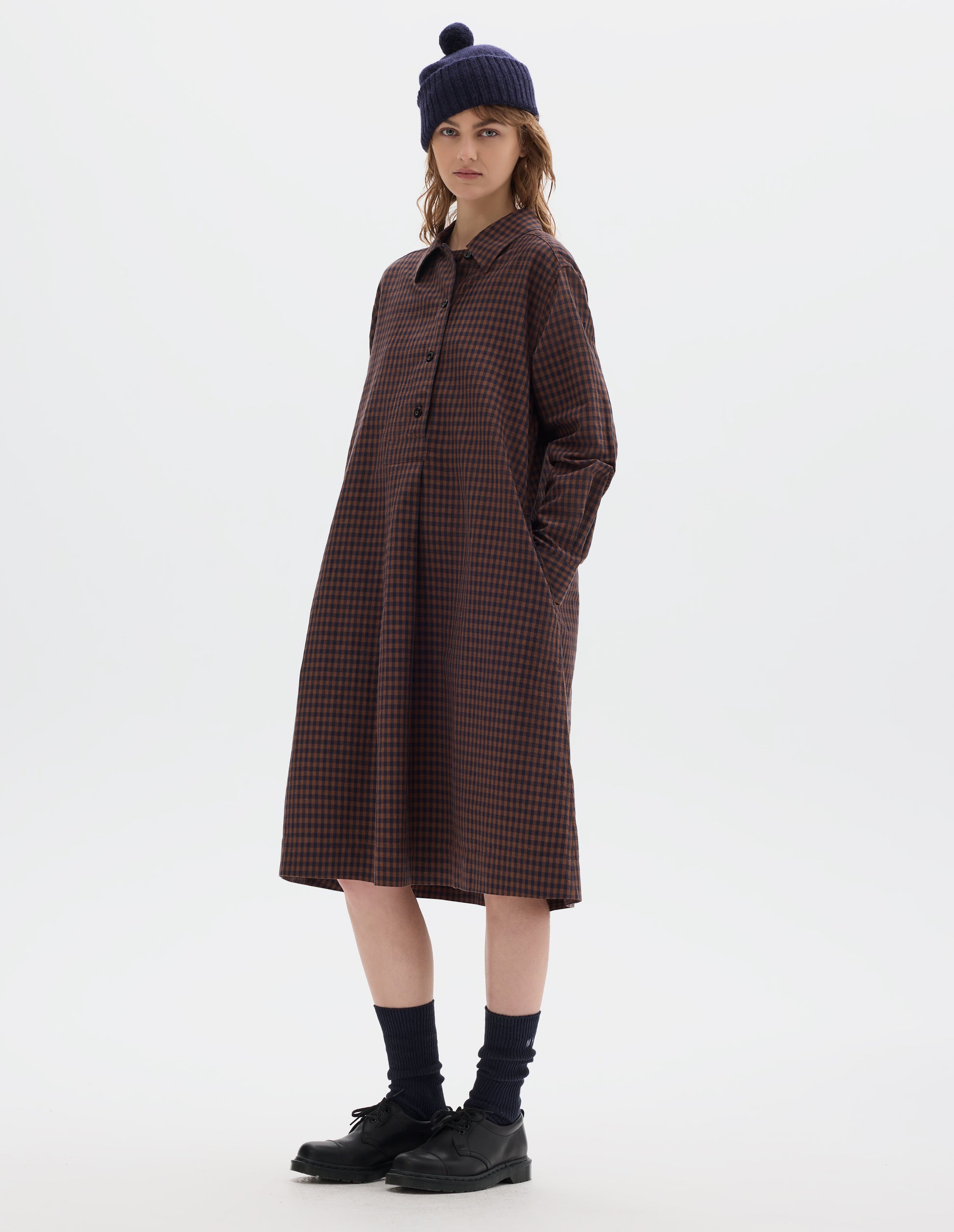 Full body image, front view of model wearing brown and dark blue check cotton linen dress, with four dark buttons and box pleat skirt. Styled with navy bobble hat, black socks and black leather shoes.