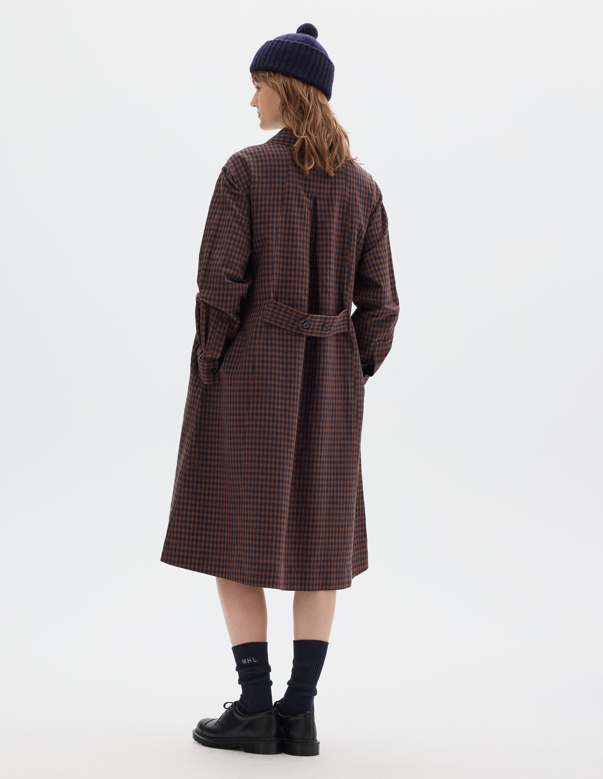 Full body image, back view of model wearing brown and dark blue check cotton linen dress, with an inverted pleat and adjustable two button belt and button cuffs. Styled with navy bobble hat, black socks and black leather shoes.